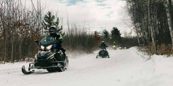 A group of people ride snowmobiles along a snow-covered trail in a forested area under a partly cloudy sky.