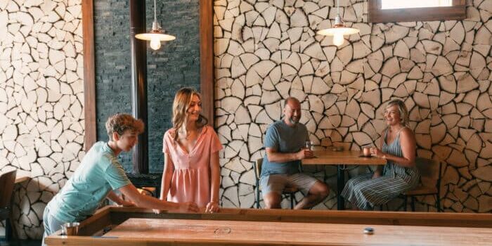 ER_Summer_Family_Dining-Craftsman-20210614_4141 Two people play shuffleboard while two others sit at a table watching and smiling in a room with stacked wood wall decor and warm lighting.