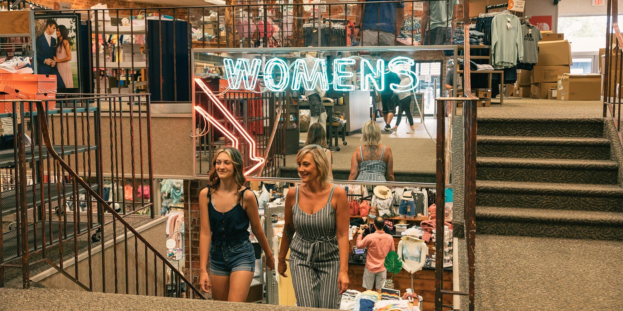 Two women walk up a carpeted staircase in a clothing store with a neon "WOMENS" sign above. Various clothing items are displayed in the background.