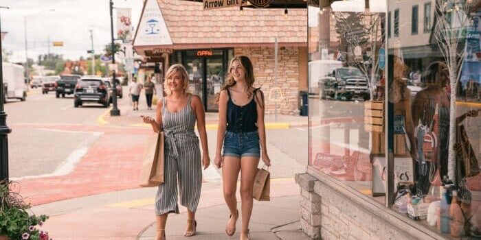 Two women walk on a sidewalk, each carrying shopping bags. They pass storefronts with displays and windows. In the background, alongside the bustling street with cars, shops, and pedestrians, there are signs for off-road routes featuring ATVs and UTVs.