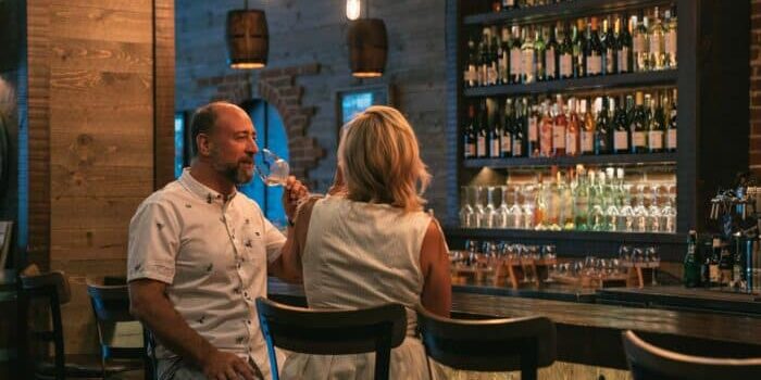 A man and a woman sit at a bar counter in a dimly lit room, drinking wine. Shelves stocked with various wine bottles are in the background, and hanging lightbulbs cast a gentle glow reminiscent of cascading waterfalls.