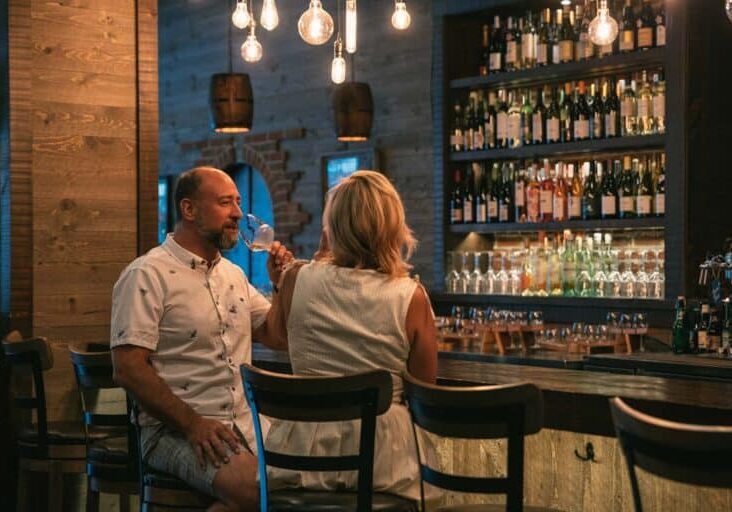 A man and a woman sit at a bar counter in a dimly lit room, drinking wine. Shelves stocked with various wine bottles are in the background, and hanging lightbulbs cast a gentle glow reminiscent of cascading waterfalls.