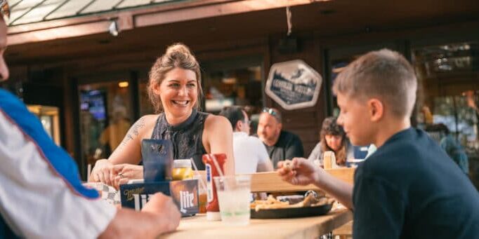 Three people sit at a wooden outdoor table eating and talking at a casual restaurant, with condiments and drinks on the table.