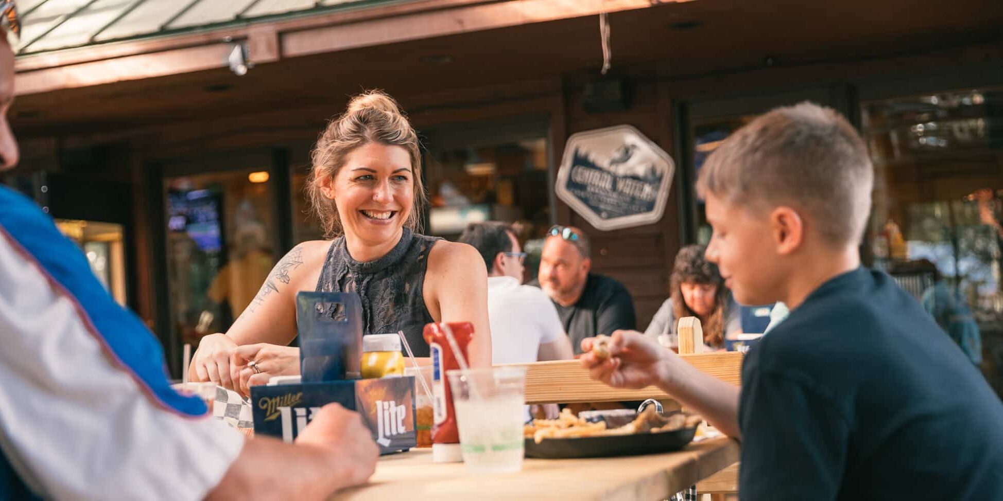 Three people sit at a wooden outdoor table eating and talking at a casual restaurant, with condiments and drinks on the table.