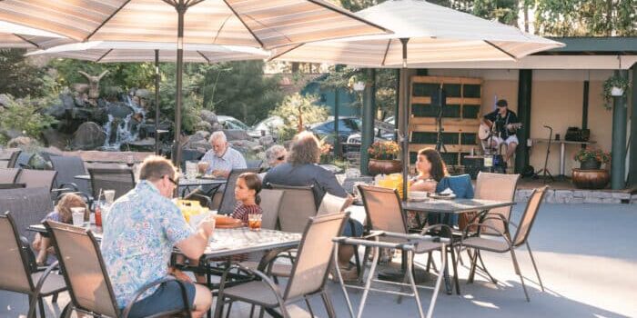 People sit at outdoor patio tables under umbrellas, eating and talking, while a musician performs on a small stage in the background amid trees and plants.