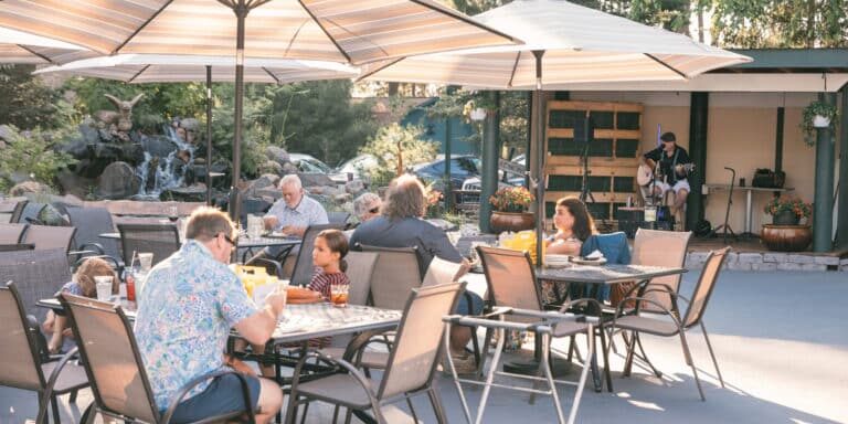 People sit at outdoor patio tables under umbrellas, eating and talking, while a musician performs on a small stage in the background amid trees and plants.