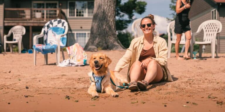 ER_WildEagleLodge_20240719_7911 A woman sits on the sand next to a golden retriever wearing a harness, with empty chairs and lakeside houses in the background.