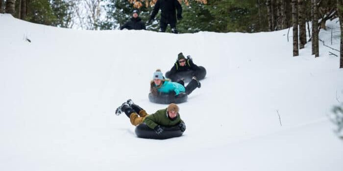 Three people wearing winter clothing are sliding down a snowy hill on inflatable tubes. A person stands at the top of the hill among trees.