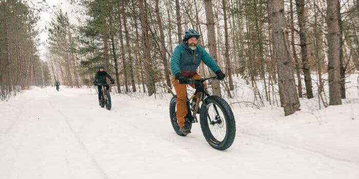 Three people ride fat-tire bikes on a snowy trail through a forest of pine trees in winter.