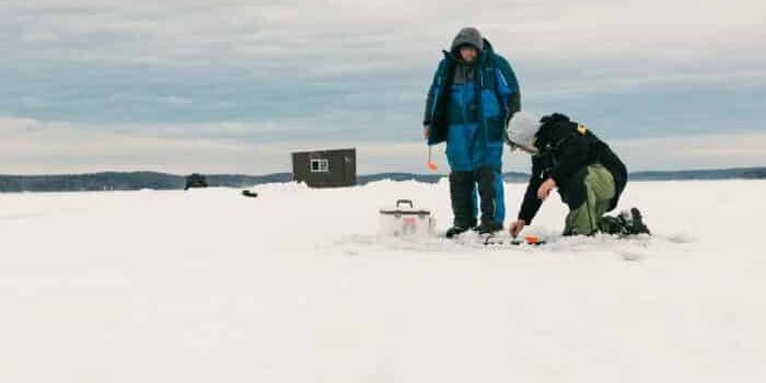 Two people are ice fishing on a snowy lake; one is kneeling by the fishing hole while the other stands nearby. An ice fishing hut and equipment are visible in the background.