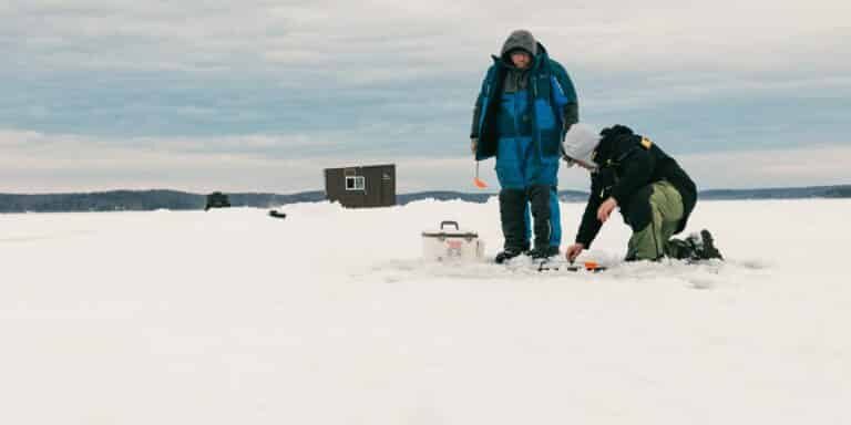 Two people are ice fishing on a snowy lake; one is kneeling by the fishing hole while the other stands nearby. An ice fishing hut and equipment are visible in the background.