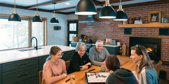 Five people sit around a wooden table in a kitchen, playing a board game and talking. Glasses of water are on the table and a fireplace is visible in the background.