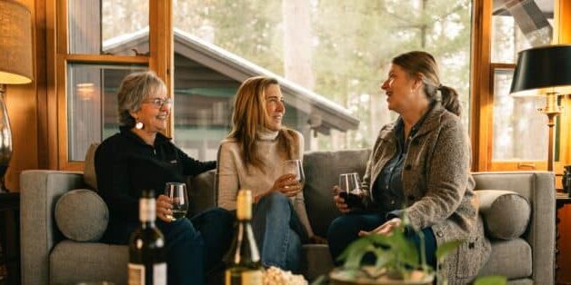 Three women sit on a couch, talking and laughing while holding drinks during their getaway. A table in front holds bottles, snacks, and plants. Large windows reveal trees, a cabin, and scenic routes outside.