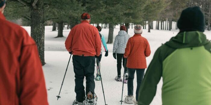 A group of people in winter clothing snowshoeing on a snowy trail surrounded by trees.