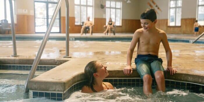 Two children talk and smile, one sitting on the edge and the other in a hot tub at an indoor pool facility. Adults sit in the background.