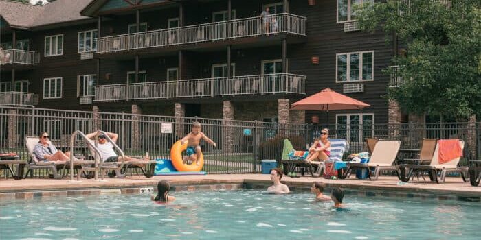 People relax by and in an outdoor pool at an apartment or resort complex; some lounge on chairs while others swim or play in the water.