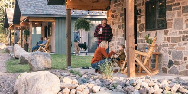 People are outside a stone house. A woman and child are on the porch, a man walks nearby, and another person sits by a building in the background.