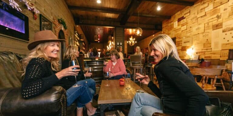 Fall_Mary_Kates_Wine_Bar_20241003_016 Several women sit in a cozy wooden lounge, chatting and drinking wine, with other people at the bar in the background.