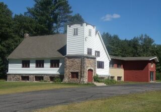 A large white house with a stone archway and chimney stands in a clearing with trees in the background, next to a red outbuilding.