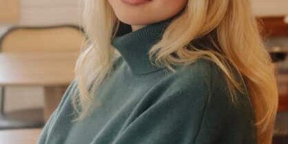 A woman with long blonde hair wearing a teal turtleneck sweater sits indoors, looking at the camera with a slight smile.