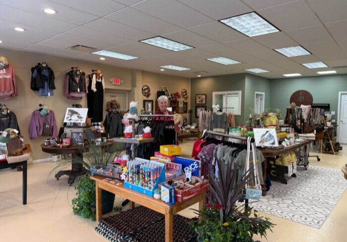 A woman stands behind a display table in a store with clothing racks, various gift items, and wall decorations in the background.