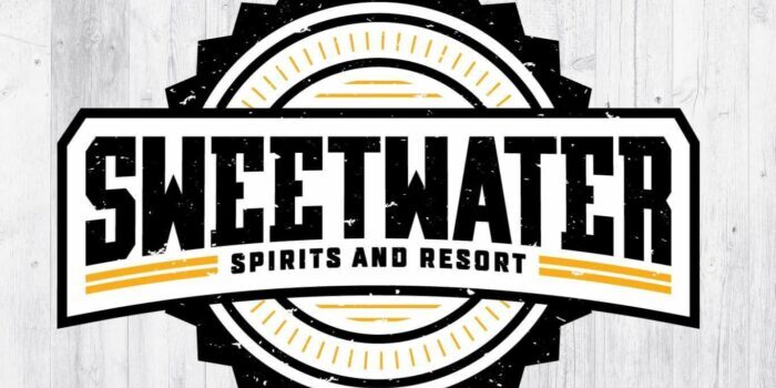 Logo of Sweetwater Spirits and Resort featuring stylized text within a circular black and yellow emblem on a wooden backdrop.