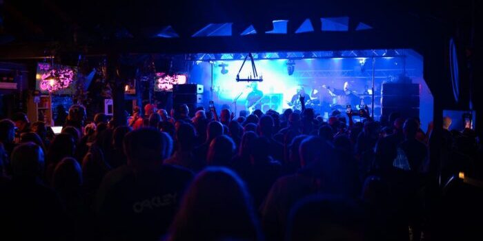 A crowd watches a live band perform on a brightly lit stage in a dark indoor venue.
