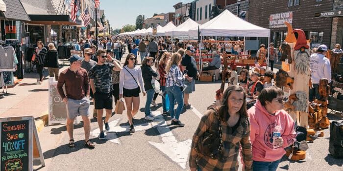 A busy outdoor market on a sunny day with people strolling past vendor tents and a variety of wooden carvings on display.