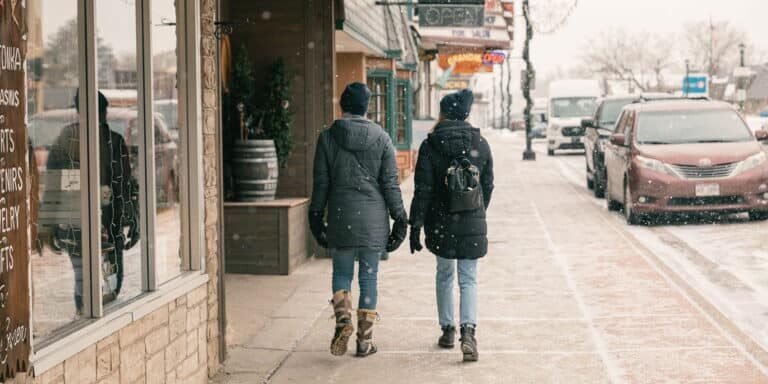 Two people wearing winter coats and backpacks walk down a snowy sidewalk in a small town. A few cars are parked on the street. Snowflakes are falling.