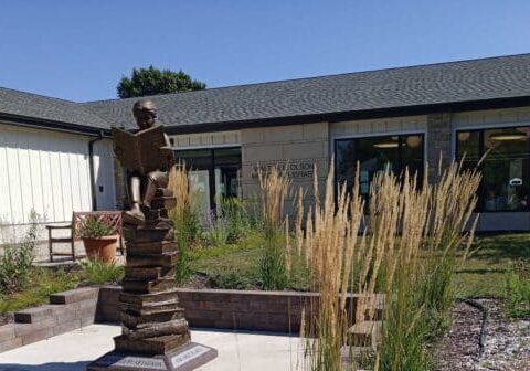 A bronze statue of a child reading a book stands in a garden outside the St. Olaf Library, capturing the spirit of learning on a sunny day.