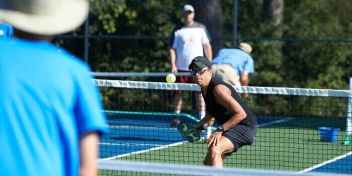 A woman wearing sunglasses and a black outfit prepares to hit a pickleball at the net during an outdoor game, with other players visible in the background.