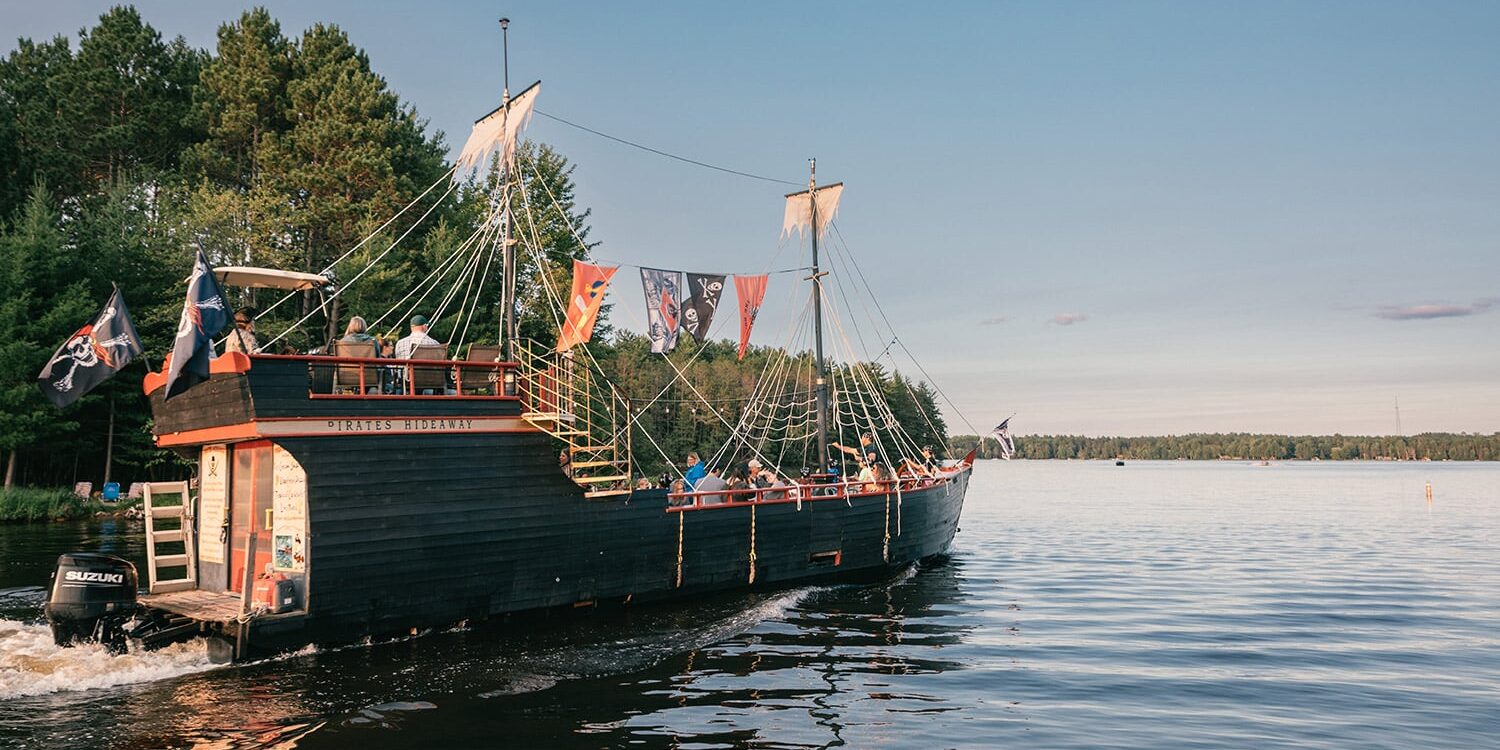 A large, black wooden ship with sails and pirate flags moves on a calm lake near a forested shoreline under a clear sky.