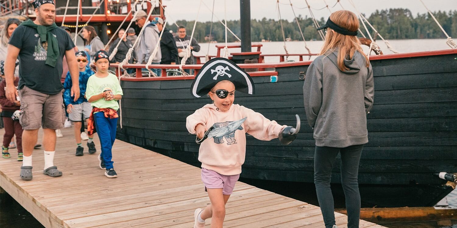 Children dressed as pirates walk and play on a wooden dock next to a large docked pirate ship, with adults and others in the background.