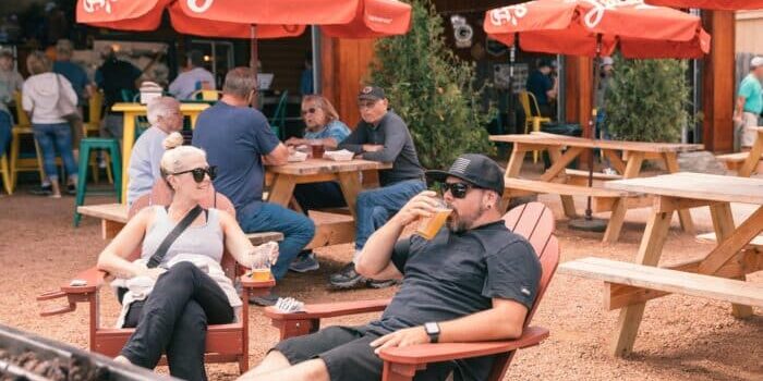 People sitting outdoors at a brewery, with red umbrellas overhead. A man drinks from a glass, while a woman holds a drink. Picnic tables are in the background.
