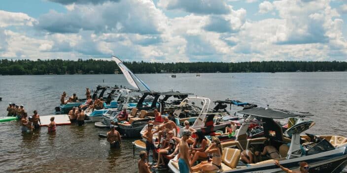 Several boats are docked side by side on a lake, with groups of people in swimsuits sitting on the boats and swimming near the shore under a partly cloudy sky.