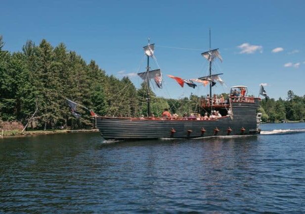 A sailing ship with tattered sails navigates a river surrounded by dense forest under a clear blue sky.