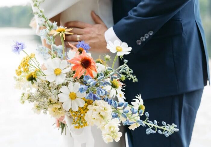 A couple stands close together outdoors, one in a white dress holding a colorful bouquet of wildflowers, and the other in a dark suit, with their arms around each other.