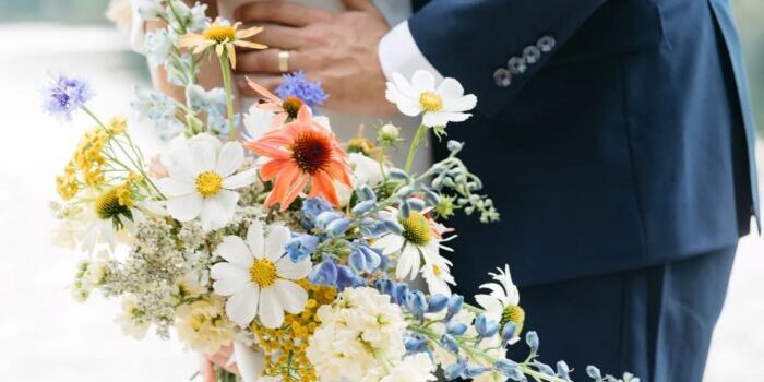 A couple stands close together outdoors, one in a white dress holding a colorful bouquet of wildflowers, and the other in a dark suit, with their arms around each other.