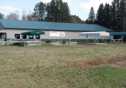 A single-story green-roofed building with canopies, picnic tables, and a sign reading "Camp." Surrounded by trees and grassy area.