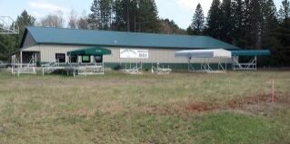 A single-story green-roofed building with canopies, picnic tables, and a sign reading "Camp." Surrounded by trees and grassy area.