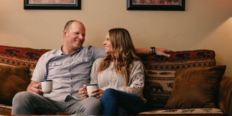 A man and woman sit on a patterned sofa holding mugs, smiling at each other, with framed artwork on the wall behind them.