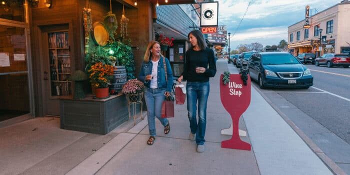 Two women walking on a sidewalk, each holding a drink. They pass a storefront with a wine sign and potted plants. Cars are parked along the street.