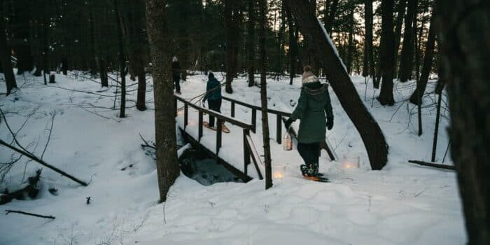 Winter_Snowshoeing_20220125_8180-1 Two people in winter coats and snowshoes cross a small wooden bridge in a snowy forest, carrying lanterns in the late afternoon light.
