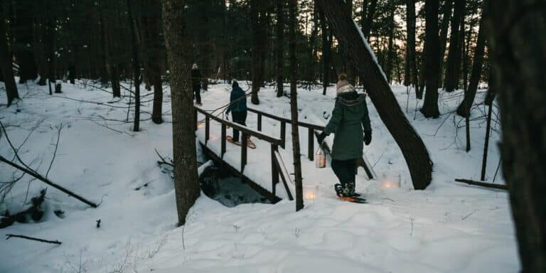 Winter_Snowshoeing_20220125_8180-1 Two people in winter coats and snowshoes cross a small wooden bridge in a snowy forest, carrying lanterns in the late afternoon light.