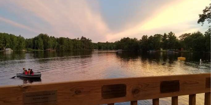 A small boat with two people on a calm lake at sunset, viewed from behind a wooden railing with plaques; trees line the shore in the background.