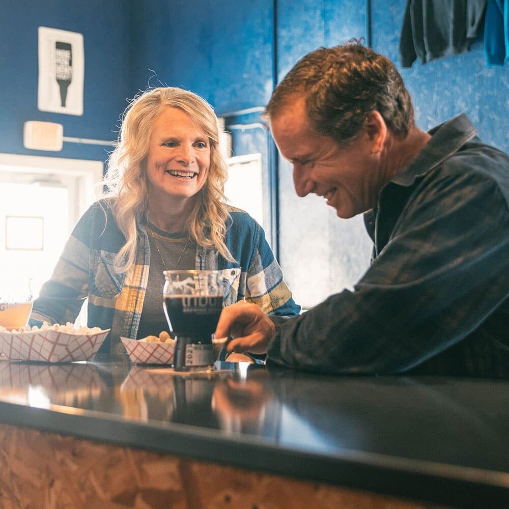 er_WinterTributeBrewery Two people sitting at a bar, laughing and talking. They have drinks and baskets of snacks in front of them, with a blue wall and Wisconsin-themed decor in the background.