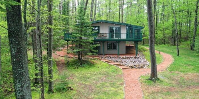 A two-story cabin surrounded by trees, with a stone path leading to the entrance, situated in a wooded area.