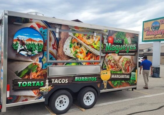 Food truck for Los Magueyes Mexican Restaurant featuring images of tortas, tacos, and burritos, parked in a lot with a man walking nearby and the restaurant sign visible.