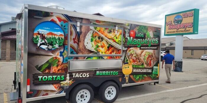Food truck for Los Magueyes Mexican Restaurant featuring images of tortas, tacos, and burritos, parked in a lot with a man walking nearby and the restaurant sign visible.