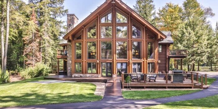 Large wooden cabin with floor-to-ceiling windows, set in a lush forest. A paved path leads to the entrance, surrounded by green grass and tall trees.